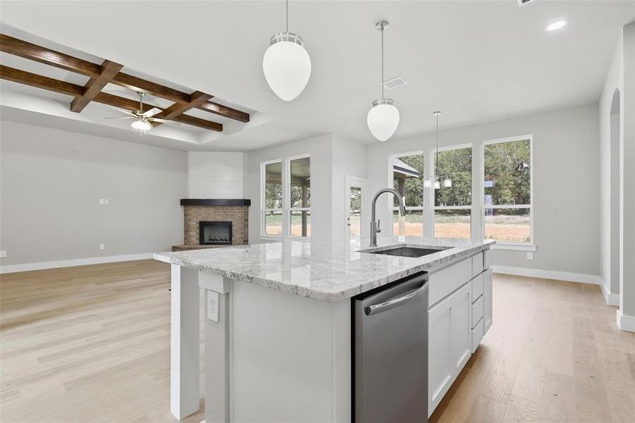 Kitchen with coffered ceiling, light stone countertops, dishwasher, an island with sink, and a fireplace