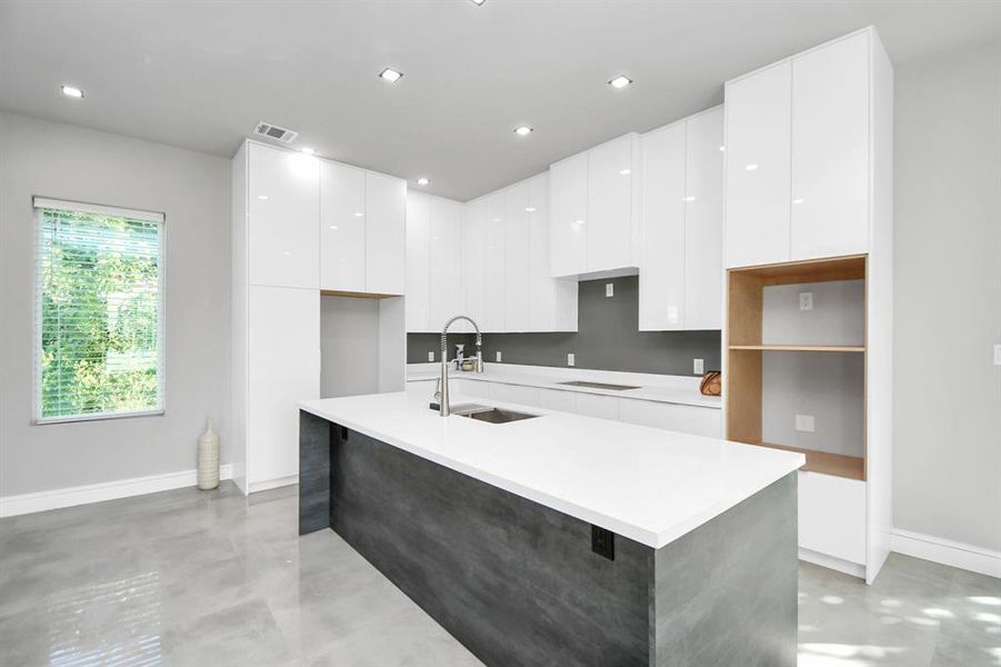 This is a modern kitchen with sleek white cabinetry, a central island with a sink, and contemporary concrete-style countertops. The space has recessed lighting and a large window providing natural light and a view of greenery.