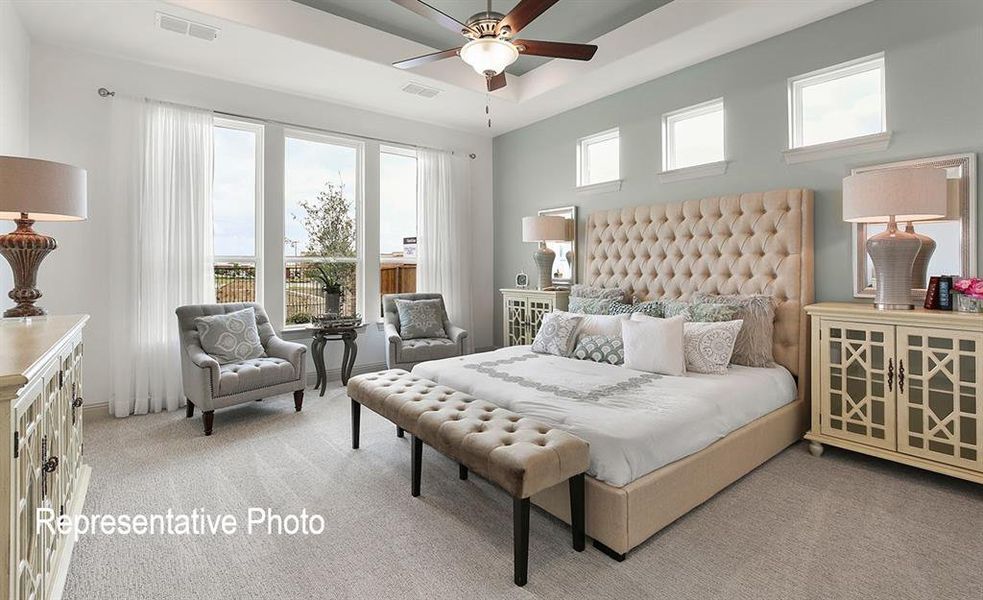 Spacious bedroom featuring light neutral carpet flooring, a tray ceiling with a fan light fixture, and a wall of three large windows providing ample natural light
