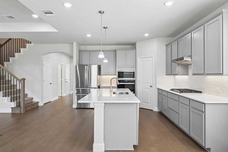 Open-concept kitchen featuring an island with sink and pendant lighting, light gray cabinetry, stainless steel appliances, and wood-finish flooring Open-concept kitchen featuring an island with sink and pendant lighting, light gray cabinetry, stainless steel appliances, and wood-finish flooring