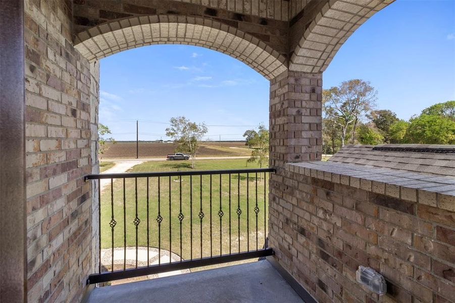 This photo shows a charming brick balcony with an arched opening and metal railing, overlooking a spacious green lawn and a scenic rural landscape.