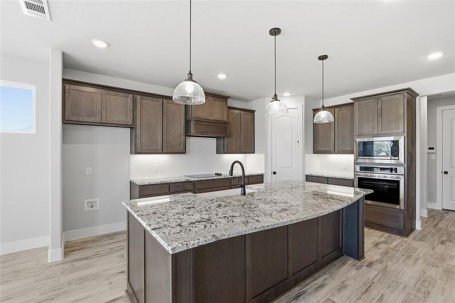 Kitchen featuring light stone countertops, light wood-style floors, appliances with stainless steel finishes, decorative light fixtures, and an island with sink