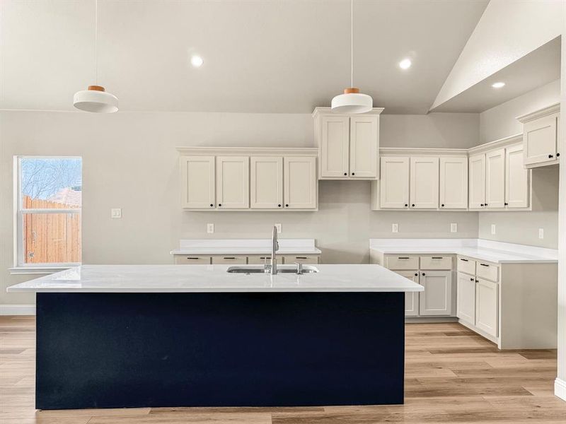 Two tone kitchen with dual tone cabinetry, a kitchen island with sink, light wood-style flooring, decorative light fixtures, and light stone countertops