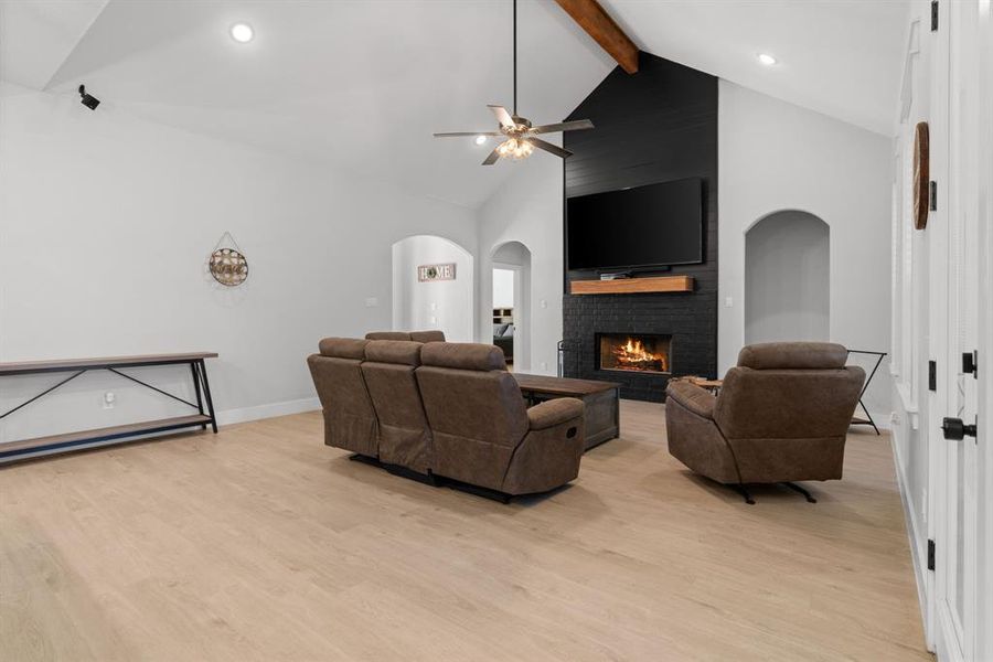 Living room featuring high vaulted ceiling, a brick fireplace, ceiling fan, beamed ceiling, and light wood-type flooring