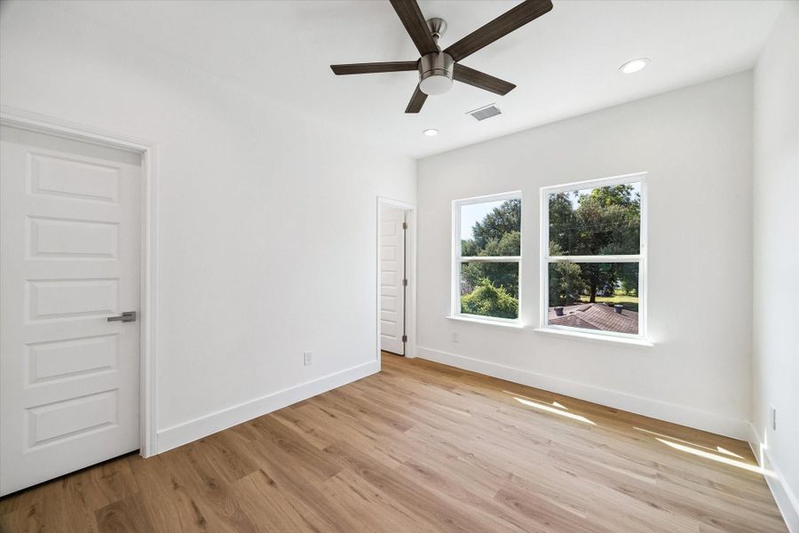 A spacious secondary bedroom is filled with natural light from two large windows overlooking mature greenery. Warm wood-look flooring, crisp white walls, recessed lighting, and a modern ceiling fan create a bright and comfortable setting. The generous floor plan provides plenty of flexibility for a full bedroom suite, desk, reading area, or additional storage pieces.