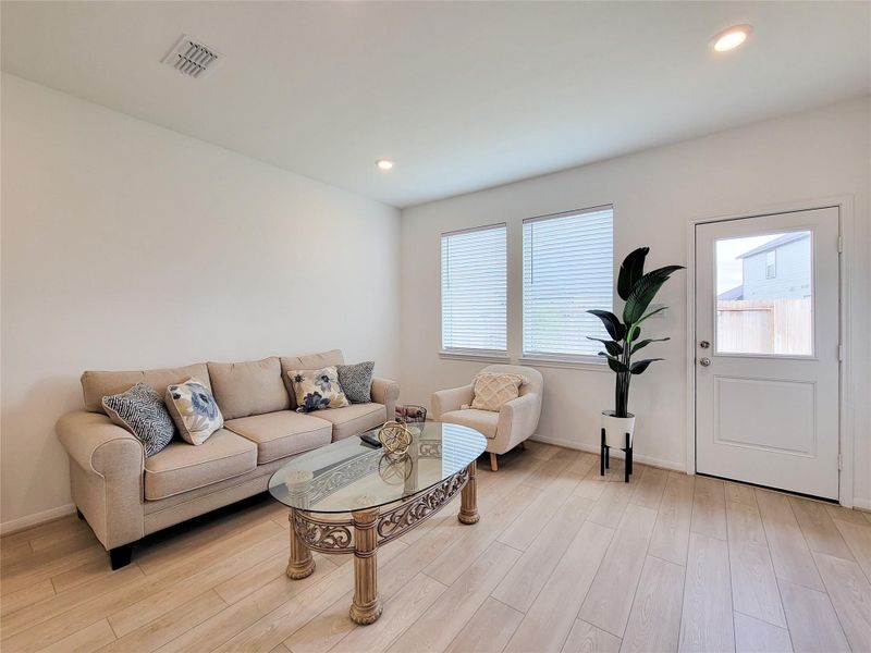 Bright and cozy living room with light wood flooring, featuring a comfortable beige sofa and armchair. The space is accented by large windows with blinds, recessed lighting.