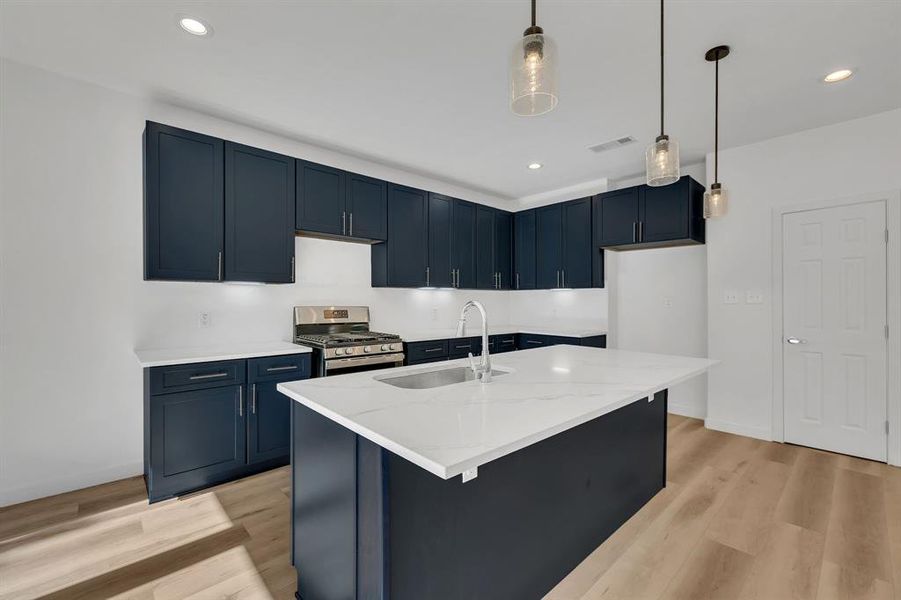 Kitchen featuring light wood-style flooring, gas stove, hanging light fixtures, a center island with sink, and light stone countertops