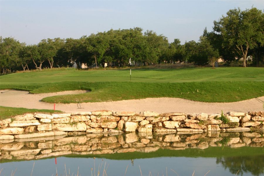 Green fairways and bunkers border a serene water feature, all framed by mature trees under an open sky. Green fairways and bunkers border a serene water feature, all framed by mature trees under an open sky.