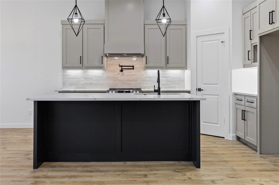 Kitchen with tasteful backsplash, light stone countertops, a center island with sink, and light wood finished floors