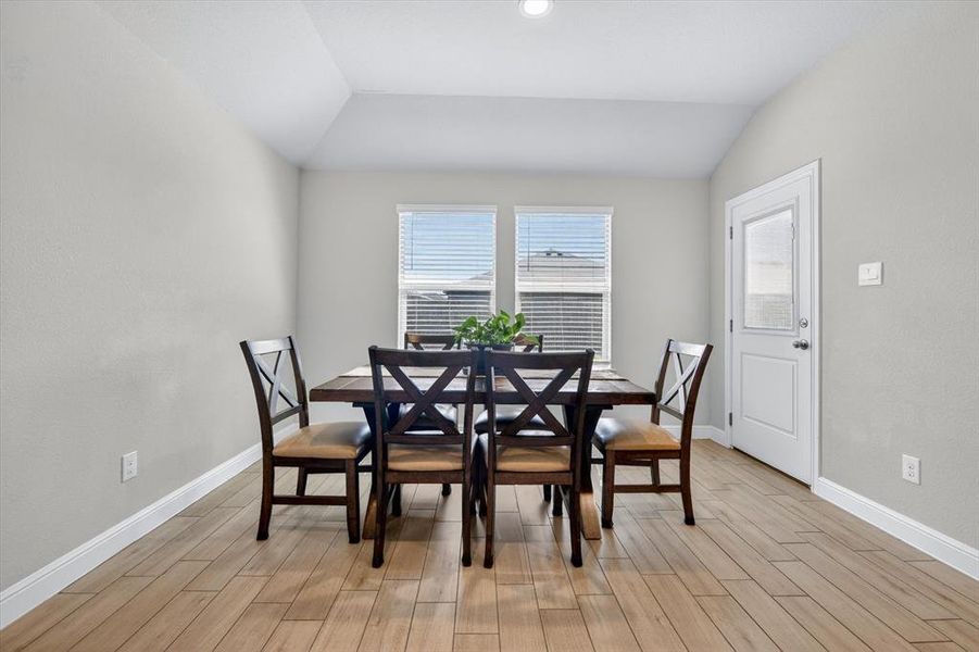 Vaulted ceiling dining area featuring wood-finish flooring, two windows with blinds, a white door with an upper glass pane, and recessed lighting