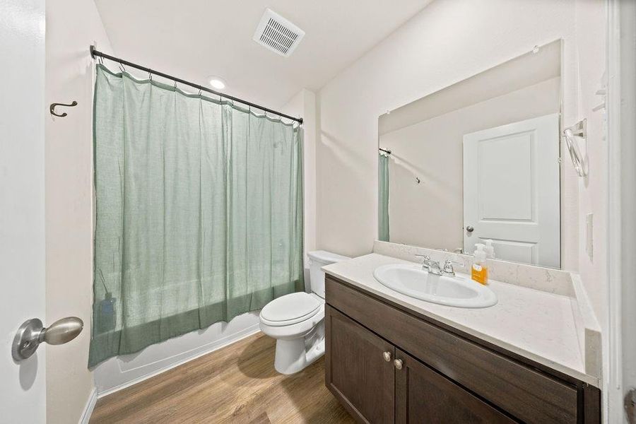 Bathroom featuring a dark wood-finish vanity with a light countertop, an undermount sink with chrome fixtures, a wall-mounted mirror, and wood-finish flooring Bathroom featuring a dark wood-finish vanity with a light countertop, an undermount sink with chrome fixtures, a wall-mounted mirror, and wood-finish flooring