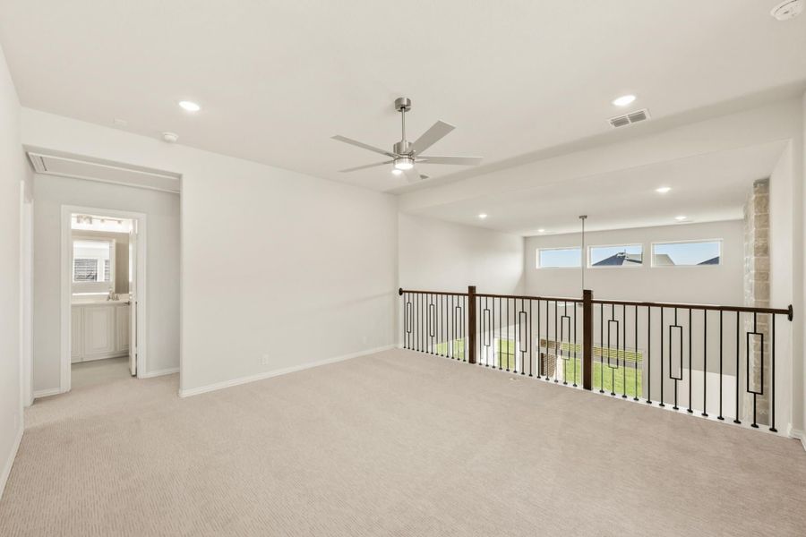Open upstairs loft with carpet, ceiling fan, iron railing overlooking lower level, and attached bathroom doorway.