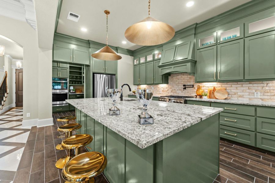 Spacious kitchen featuring a large central island with a light-colored stone countertop, two brass pendant lights, and ample green cabinetry
