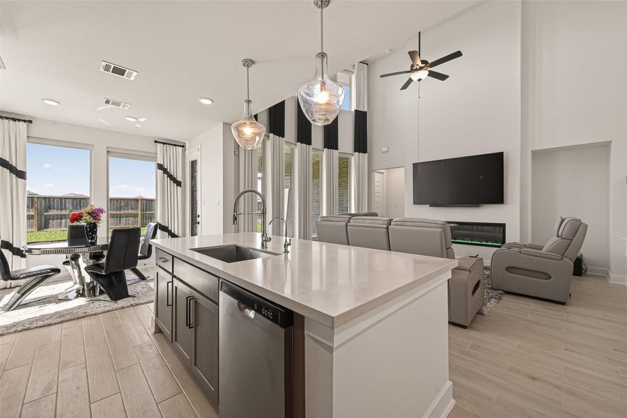 This modern open-concept living area features a spacious kitchen island with pendant lighting, sleek quartz countertops, and stainless steel appliances. The living room boasts high ceilings, a mounted TV, and comfortable seating, complemented by floor to ceiling windows that offer plenty of natural light and a view of the backyard.