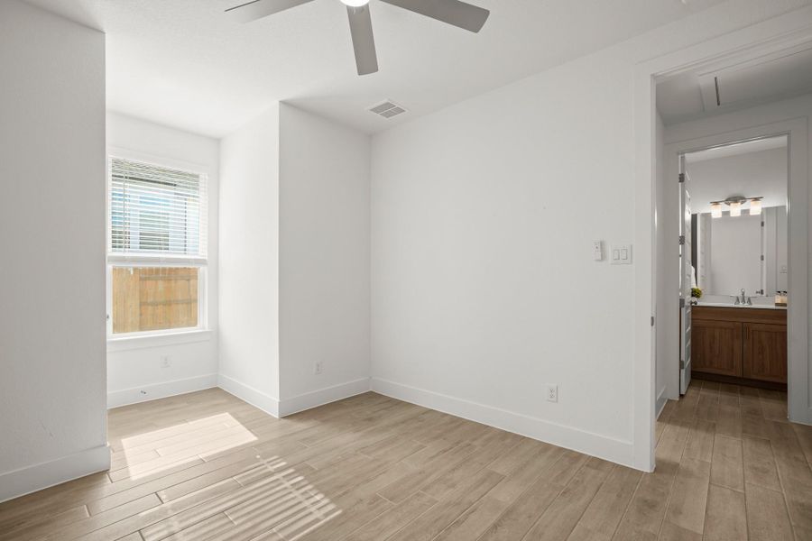 A well-lit bedroom features wood-look tile flooring and a ceiling fan, with a glimpse into a connected bathroom.
