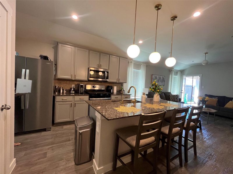 Modern kitchen with sleek gray cabinets, granite countertops, and stainless steel appliances. It features a central island with seating, pendant lighting, and a bright, open layout.