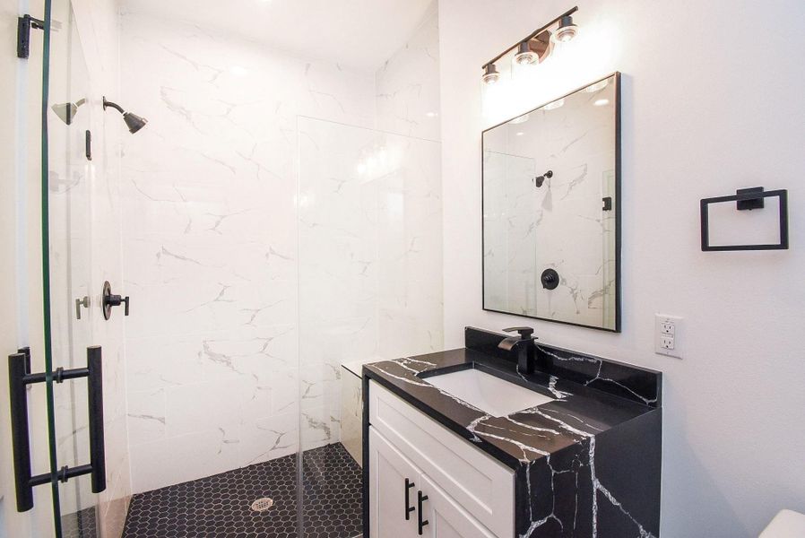 Modern bathroom with a sleek, glass-enclosed shower featuring marble-style walls and a dark hexagonal tile floor. The vanity has a black marble countertop with a white cabinet, complemented by a large mirror and stylish lighting.