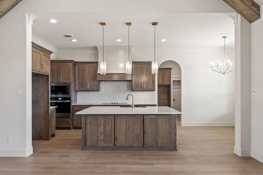 Gourmet kitchen featuring a central island with sink, wood-finish cabinetry, built-in double ovens, and a tile backsplash