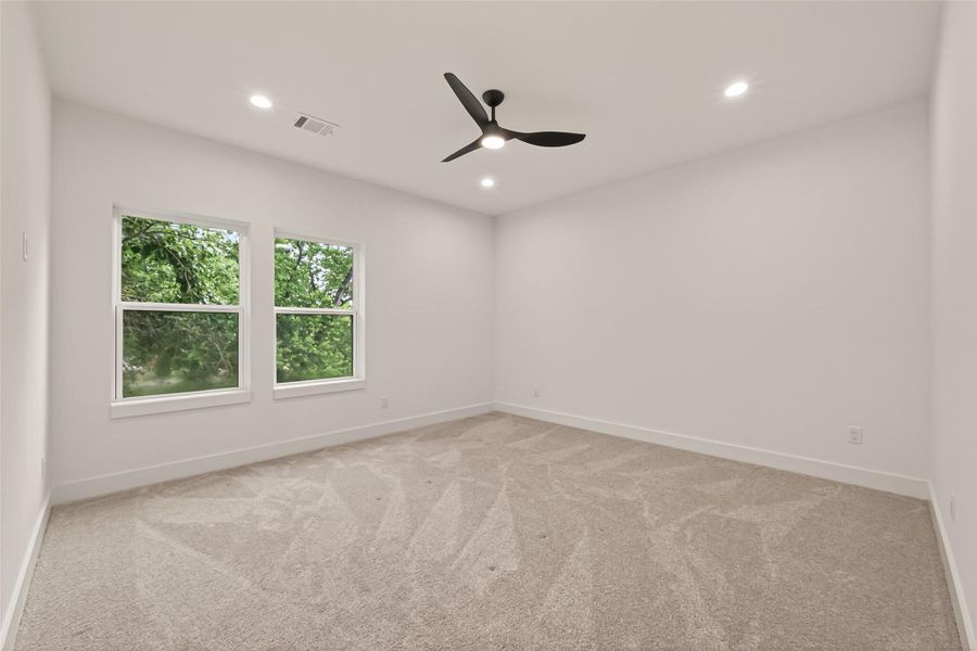 This room features clean white walls, a modern ceiling fan, and plush carpeting. Natural light streams through two large windows, offering a view of lush greenery, creating a bright and inviting space. This room features clean white walls, a modern ceiling fan, and plush carpeting. Natural light streams through two large windows, offering a view of lush greenery, creating a bright and inviting space.