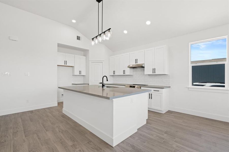 Modern kitchen with white cabinetry, stainless steel appliances, an island with a sink, and stylish pendant lighting. Features wood flooring and a bright, airy feel with a window for natural light.