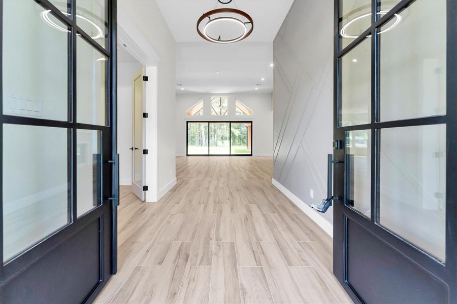 Entryway with elegant glass doors and sleek, light wood flooring. The spacious hallway leads to the living room with large windows, offering plenty of natural light and a view of greenery outside.