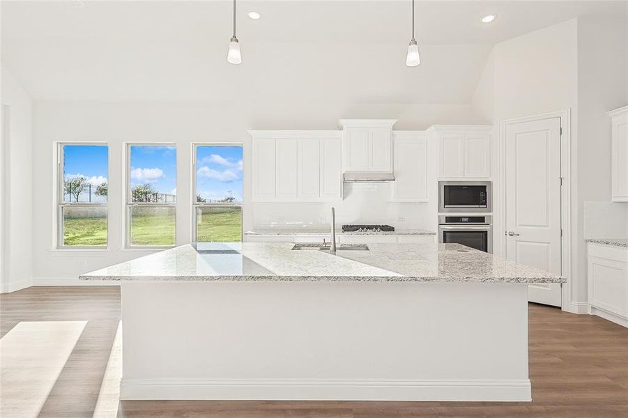 Kitchen featuring light stone counters, an island with sink, white cabinetry, stainless steel appliances, and decorative light fixtures