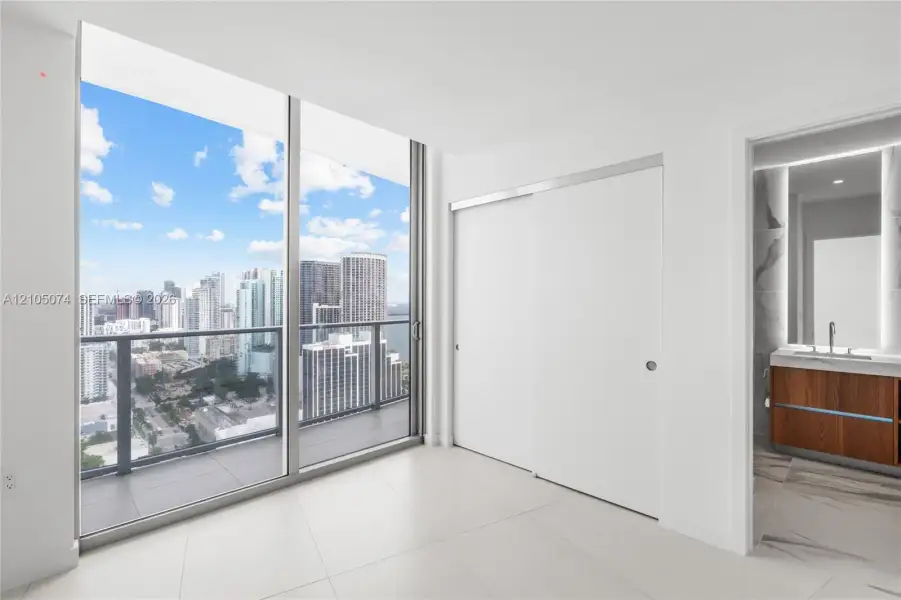 Floor-to-ceiling windows, private balcony with metal railing, sliding closet doors, wood-finish vanity with countertop basin, and stone-finish flooring