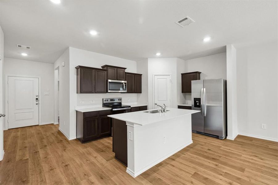 Kitchen featuring wood-finish flooring, white countertops, dark wood cabinetry, a central island with an integrated sink, and stainless steel appliances