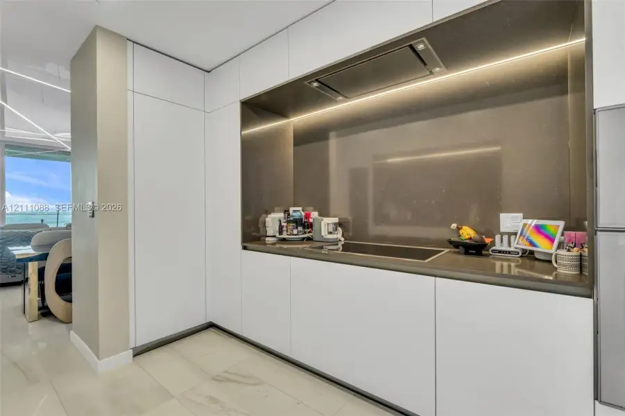 Sleek kitchen featuring matte white flat-panel cabinetry, a recessed cooktop, integrated range hood, solid-surface backsplash, and polished stone-finish flooring Sleek kitchen featuring matte white flat-panel cabinetry, a recessed cooktop, integrated range hood, solid-surface backsplash, and polished stone-finish flooring