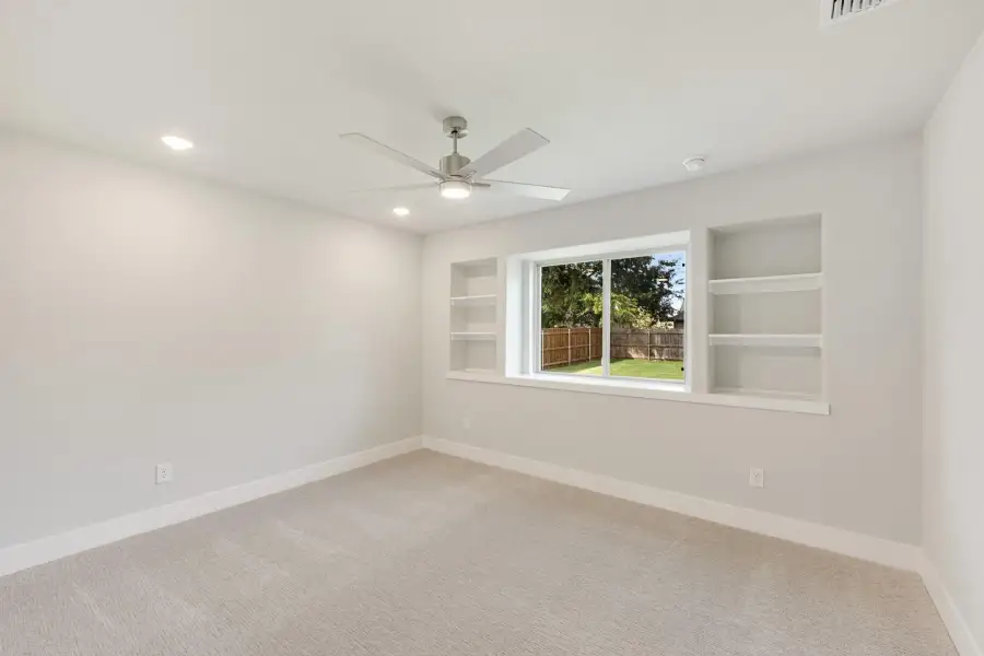 Empty bedroom with carpet, ceiling fan, built-in shelves, and a window overlooking a fenced backyard