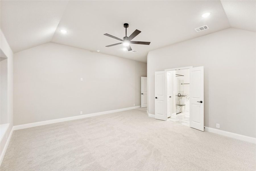 Spacious bedroom with vaulted ceiling, ceiling fan, carpet flooring, and open doors to ensuite bathroom