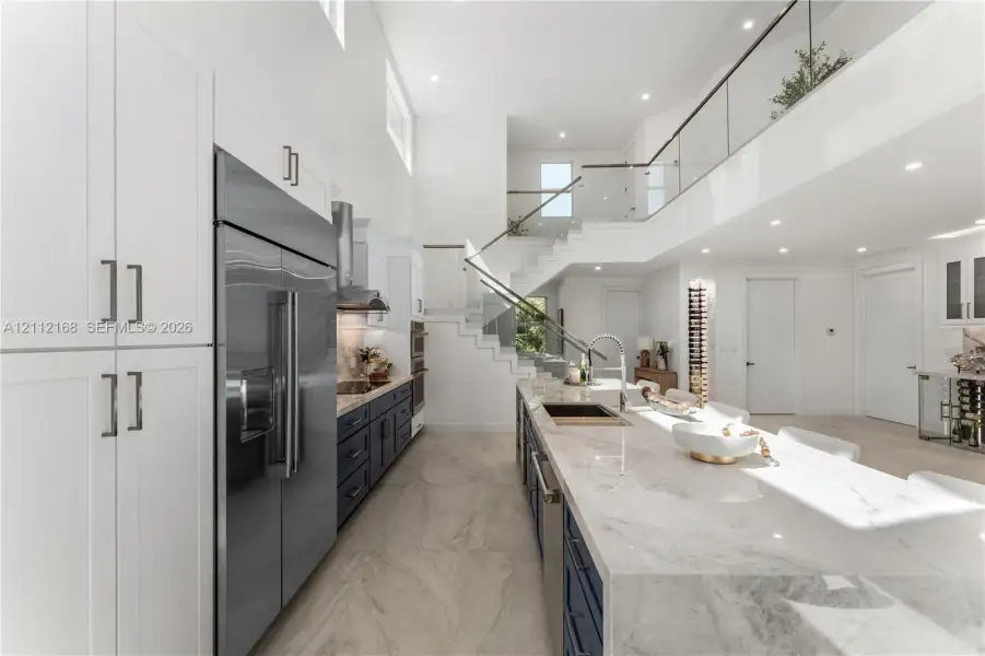 High-ceilinged kitchen featuring dual-tone cabinetry, a waterfall-edge stone island, recessed lighting, and a glass-panel staircase railing High-ceilinged kitchen featuring dual-tone cabinetry, a waterfall-edge stone island, recessed lighting, and a glass-panel staircase railing