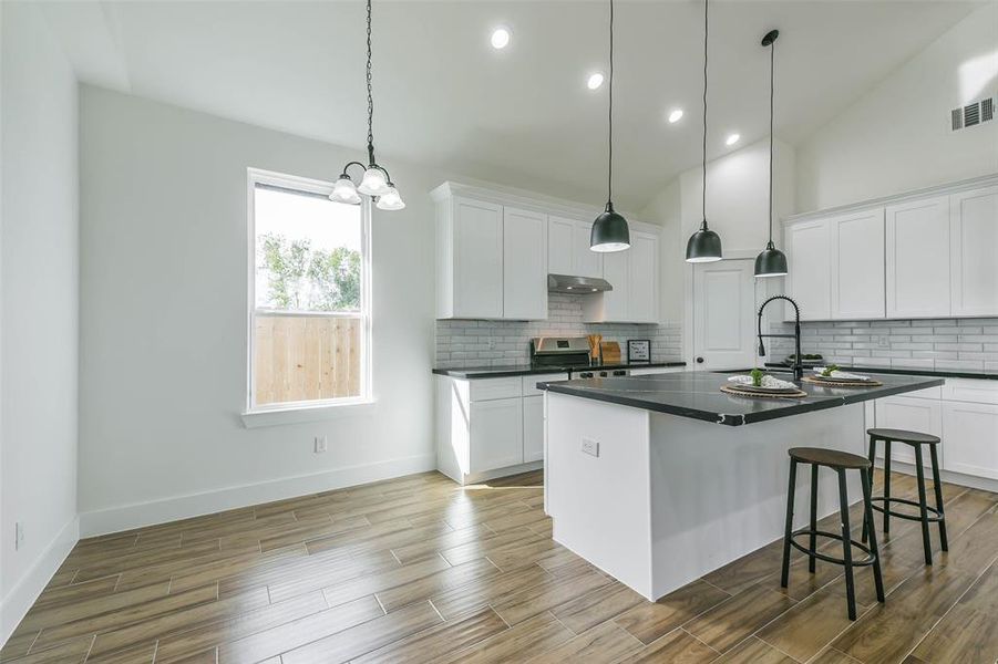 This is a bright, modern kitchen with white cabinetry, stainless steel appliances, and a central island with bar seating. It features elegant pendant lighting and a stylish subway tile backsplash. The space is complemented by wood-look flooring and has a large window for natural light.