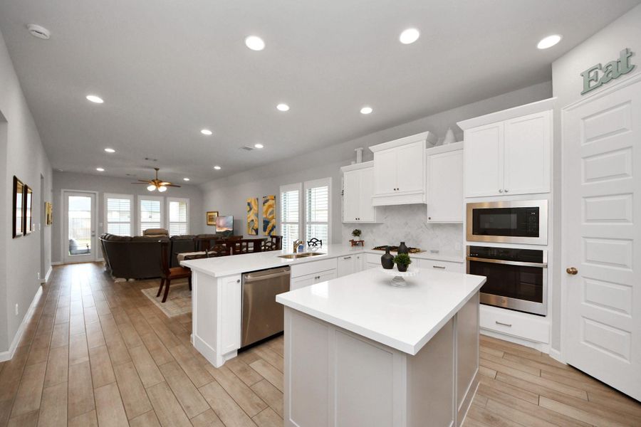 This spacious, open-concept kitchen and living area features modern white cabinetry, stainless steel appliances, and a central island. The room is bright and airy, with recessed lighting and large windows, leading into a cozy living space with a ceiling fan and wooden flooring. This spacious, open-concept kitchen and living area features modern white cabinetry, stainless steel appliances, and a central island. The room is bright and airy, with recessed lighting and large windows, leading into a cozy living space with a ceiling fan and wooden flooring.