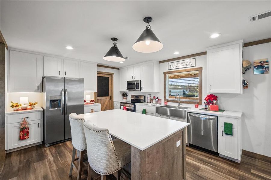 Modern kitchen featuring a large central island with a solid surface countertop, white shaker-style cabinetry, stainless steel appliances, and wood-finish flooring