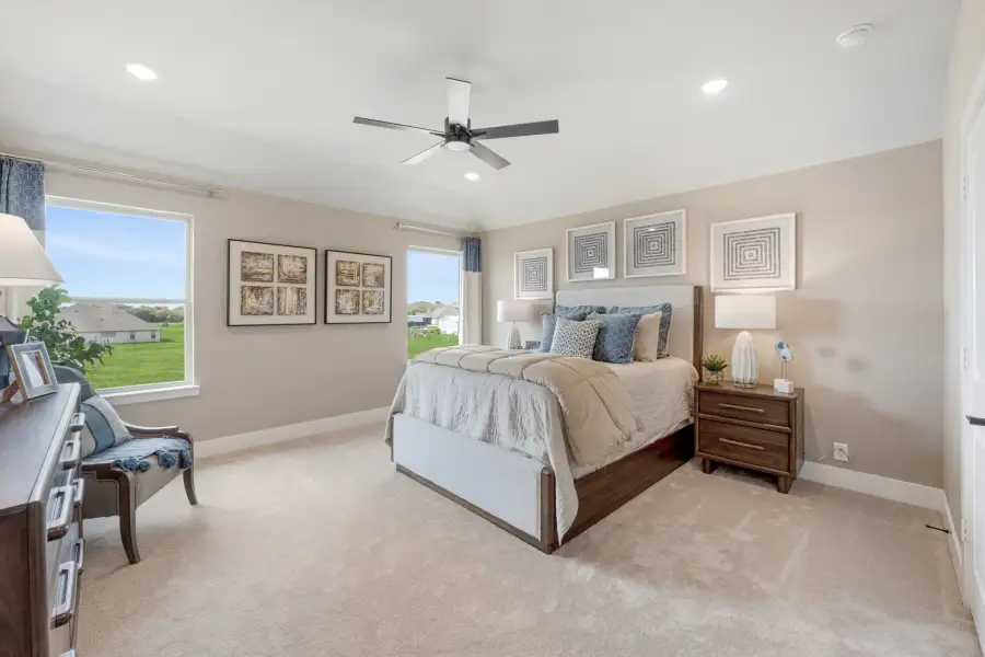 Spacious bedroom with wood bed frame, ceiling fan, carpet flooring, and two large windows with outdoor views. Spacious bedroom with wood bed frame, ceiling fan, carpet flooring, and two large windows with outdoor views.
