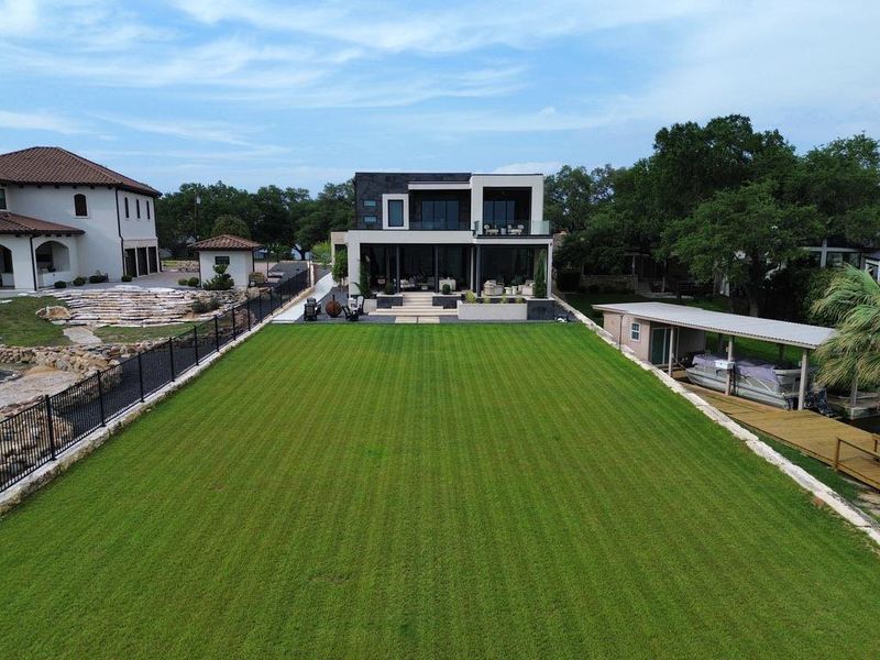 Expansive green lawn extending towards a modern multi-story residence with a boat dock and covered boat lift