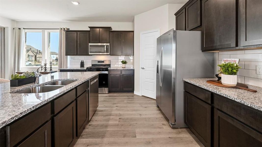 Kitchen with dark brown cabinetry, stainless steel appliances, light wood-type flooring, light stone counters, and recessed lighting Kitchen with dark brown cabinetry, stainless steel appliances, light wood-type flooring, light stone counters, and recessed lighting