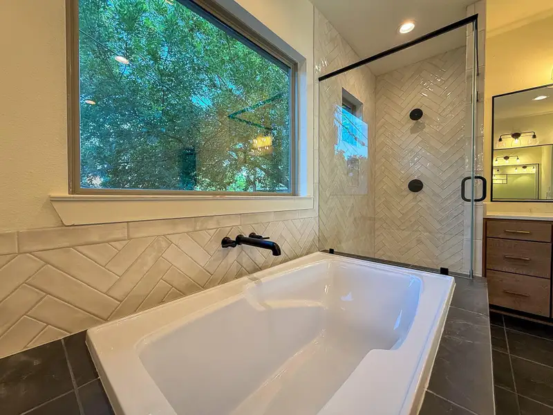 A luxurious, deep soaking tub with breathtaking natural light is flanked by a stunning, glass-enclosed shower adorned with intricate, floor to ceiling herringbone tile.