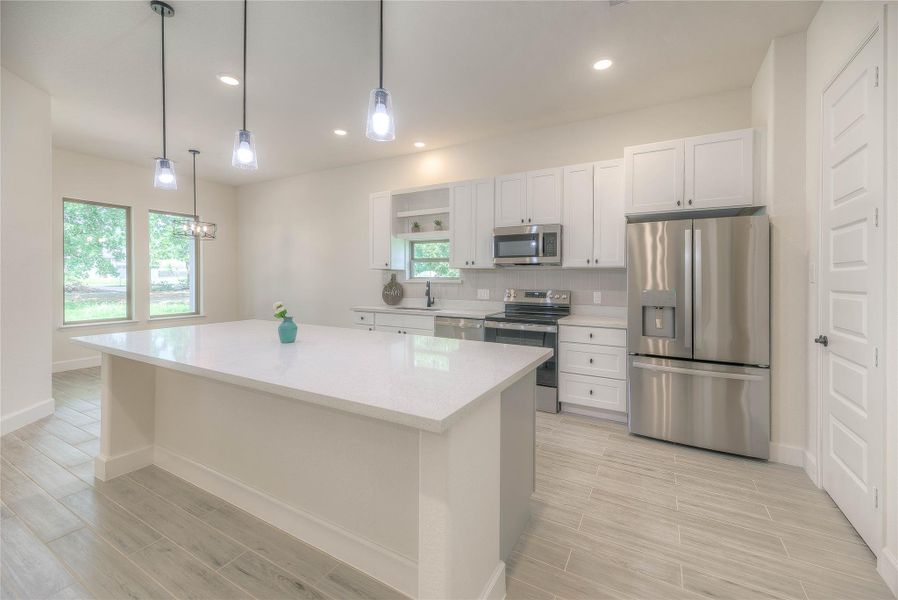 This modern kitchen features sleek stainless steel appliances, ample white cabinetry, and a spacious island with pendant lighting. Large windows provide natural light, and the light wood flooring adds warmth to the space.