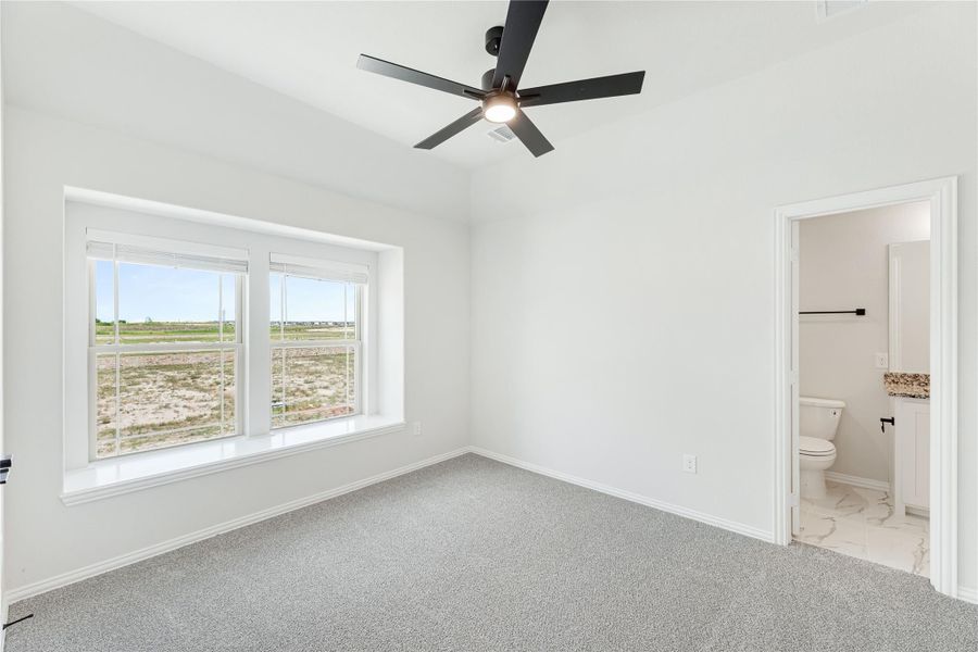 Bedroom with gray carpet, black ceiling fan, large window, and ensuite bathroom doorway visible