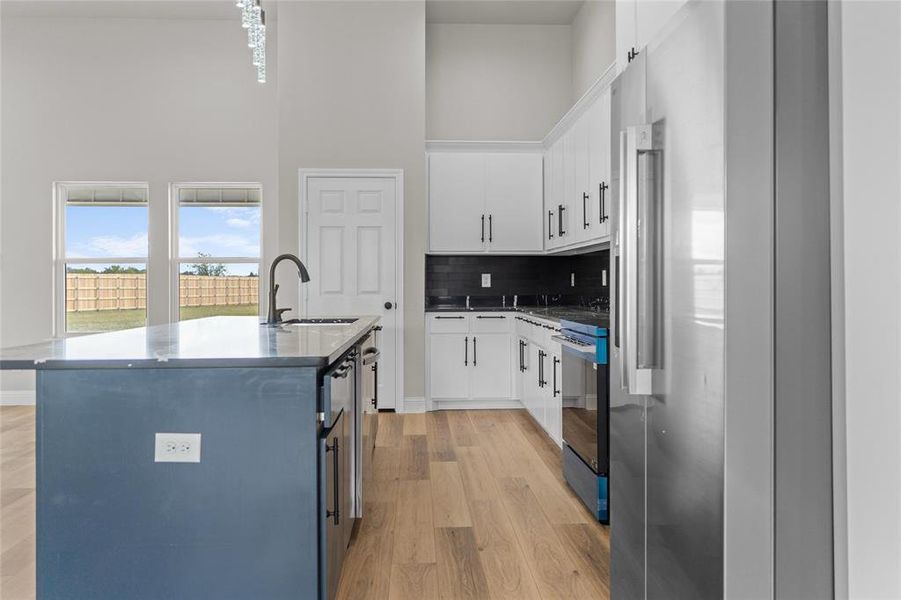 Kitchen with white cabinetry, stainless steel refrigerator, a kitchen island with sink, tasteful backsplash, and a towering ceiling