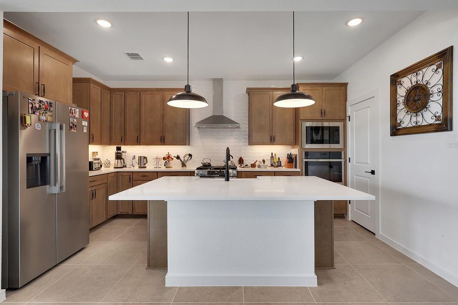Modern kitchen with warm wood cabinetry, stainless steel vent hood and appliances, and a large quartz island with sink and dishwasher. Features under-cabinet lighting, pendant lights, and a built-in oven and microwave. Ideal for cooking and entertaining.
