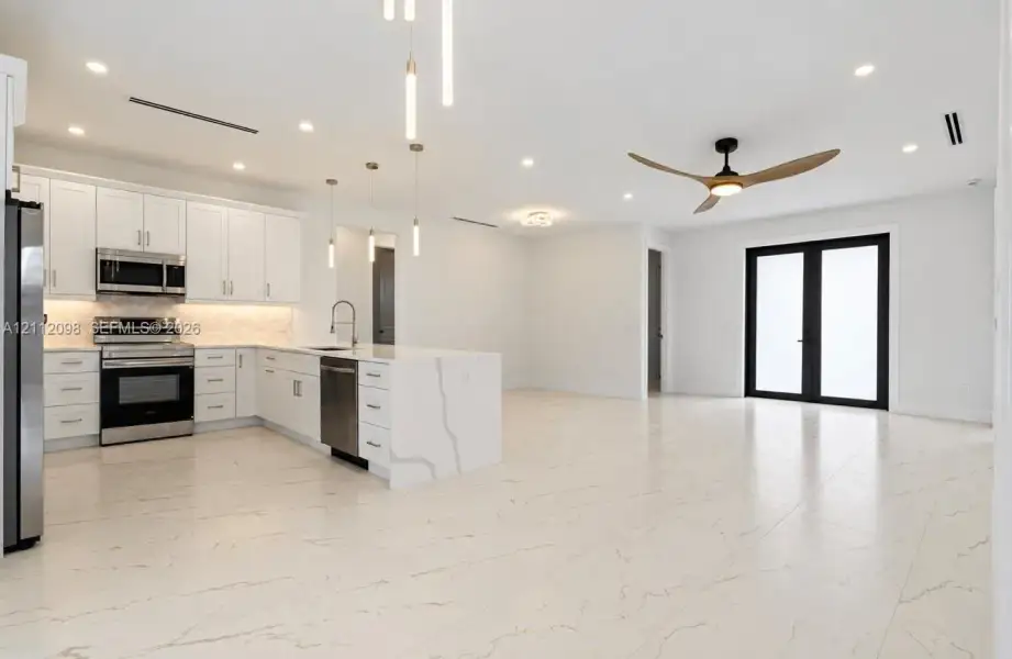 Kitchen featuring white shaker-style cabinetry, an expansive island with waterfall edge, integrated stainless steel appliances, recessed ceiling lighting, and large-format tile flooring Kitchen featuring white shaker-style cabinetry, an expansive island with waterfall edge, integrated stainless steel appliances, recessed ceiling lighting, and large-format tile flooring