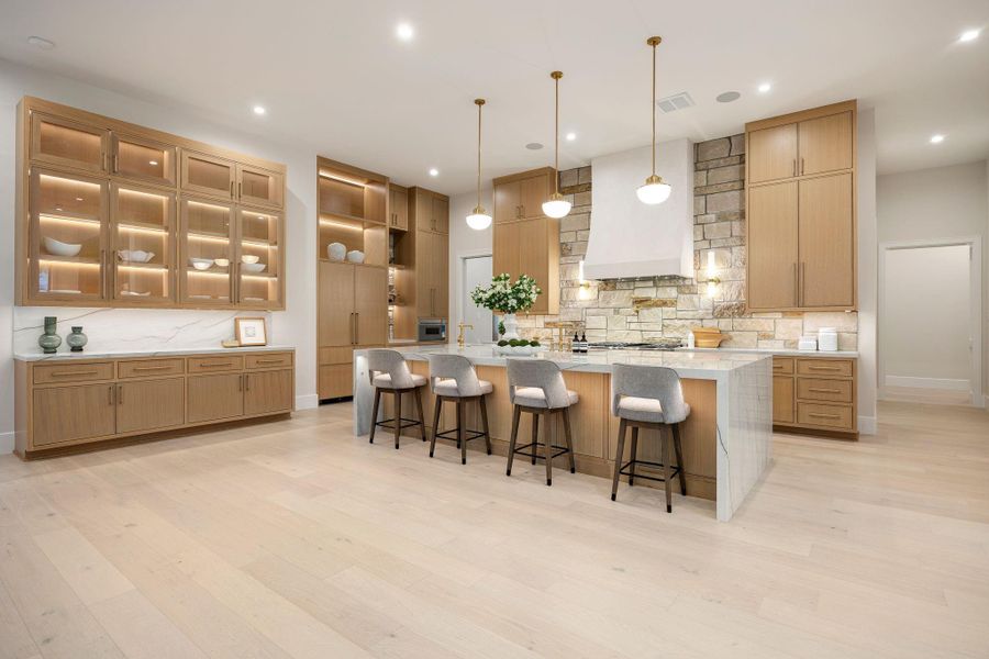 A beautifully crafted kitchen featuring custom micro-shaker cabinetry, warm finishes, and a large central island designed for gathering and entertaining . Thoughtful details like a pot filler and dedicated pantry space enhance everyday functionality, while layered lighting and clean lines create a timeless aesthetic. The open layout connects seamlessly to the living areas, making it the heart of the home.