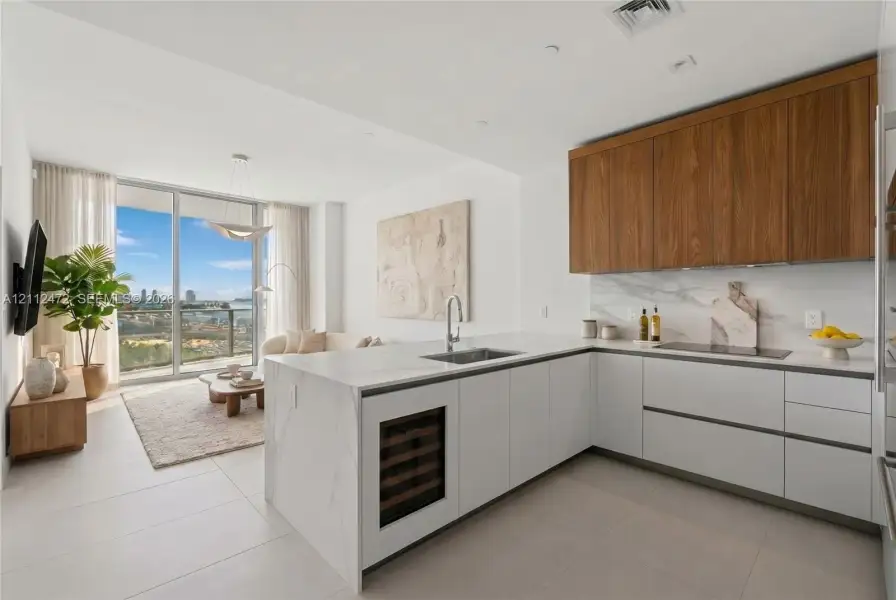 Kitchen featuring custom wood-veneer upper cabinetry, matte white lower cabinets, stone-surfaced island with an integrated wine refrigerator, waterfall-edge countertop, and full-height stone backsplash. VIRTUALLY STAGED