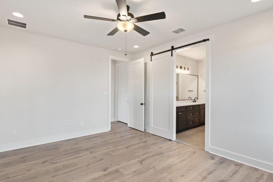 Unfurnished bedroom featuring recessed lighting, light wood-style flooring, a barn door, a ceiling fan, and ensuite bath