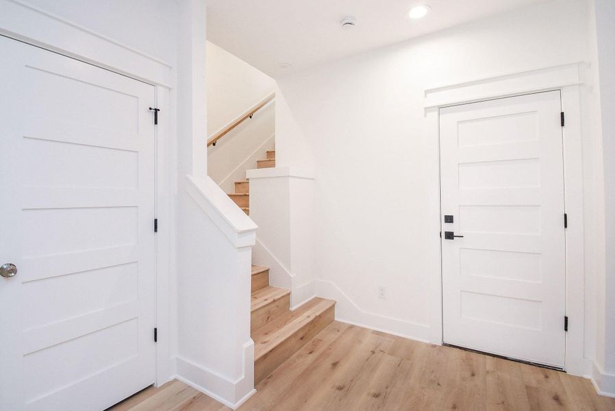 This photo showcases a bright and modern entryway with light wood flooring and crisp white walls. It features a staircase with wooden steps and white railings, along with two paneled doors. The space is well-lit with recessed lighting, offering a clean and welcoming impression.