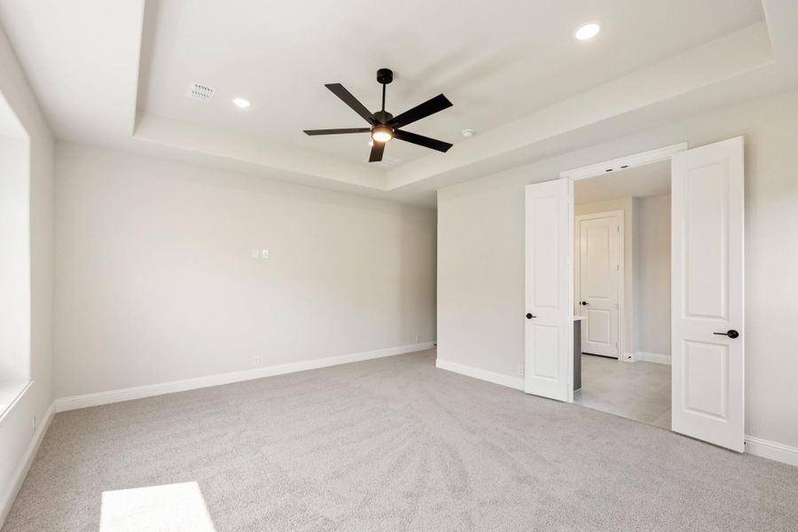 Empty bedroom with tray ceiling, black ceiling fan, gray carpet, and white double doors leading to hallway
