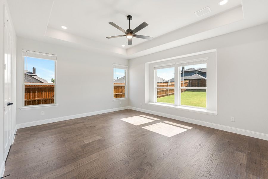 Empty room with tray ceiling, ceiling fan, dark hardwood floors, and three windows overlooking a backyard Empty room with tray ceiling, ceiling fan, dark hardwood floors, and three windows overlooking a backyard